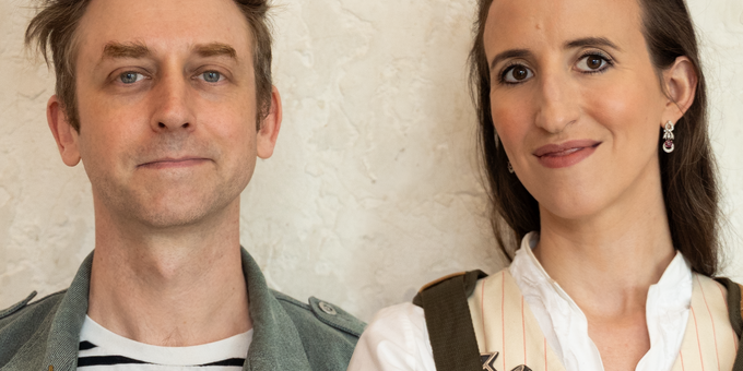 Two people wearing leather and canvas aprons, photographed against a textured light wall. On the left, a man with tousled brown hair in a striped shirt and denim vest with a small enamel pin. On the right, a woman with dark hair pulled back, wearing a white striped blouse with a metal brooch and drop earrings.